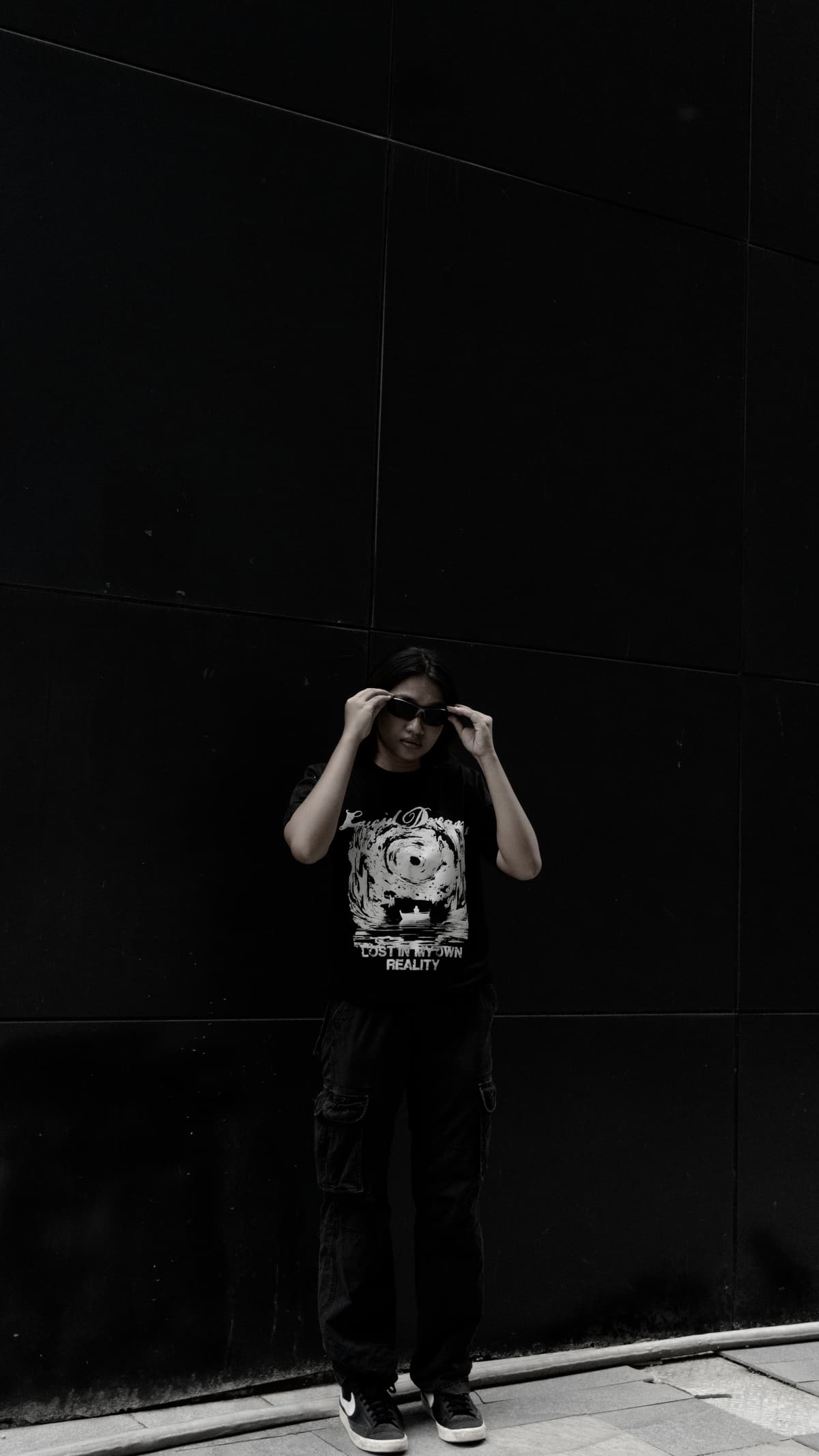 A model in the LD Signature Shirt in Black, adjusting her sunglasses against a dark panelled wall